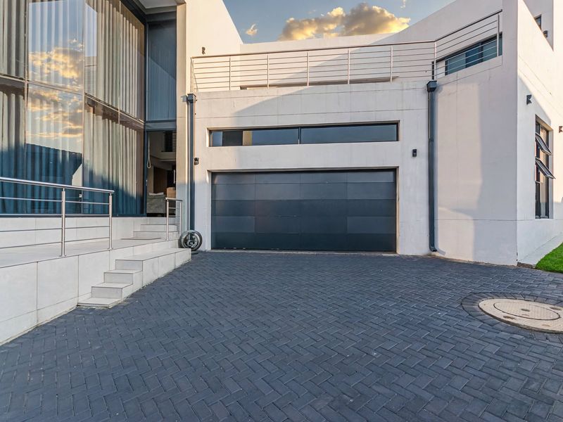 Modern architectural front elevation featuring a dark garage door, paved driveway, and a striking glass facade with an elevated balcony.