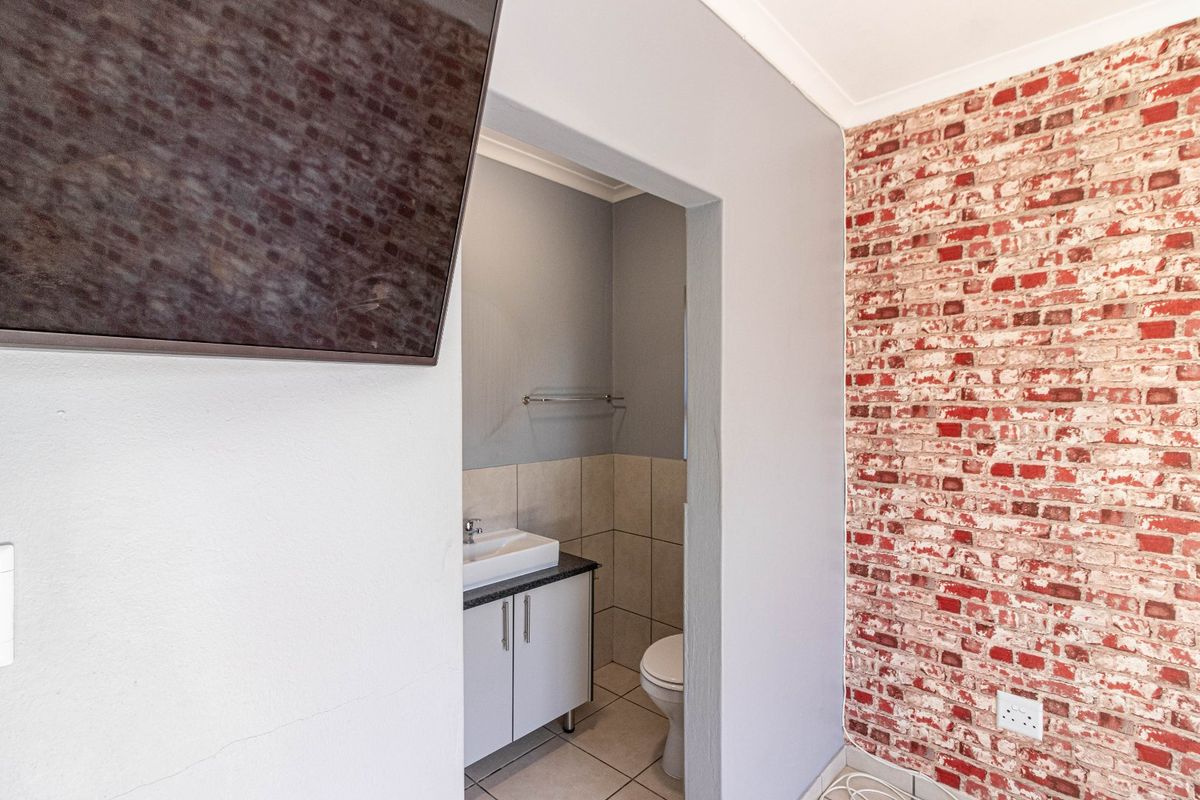 A compact bathroom featuring a vanity with basin, toilet, and tiled finishes, viewed from an adjacent room with a brick-effect feature wall.