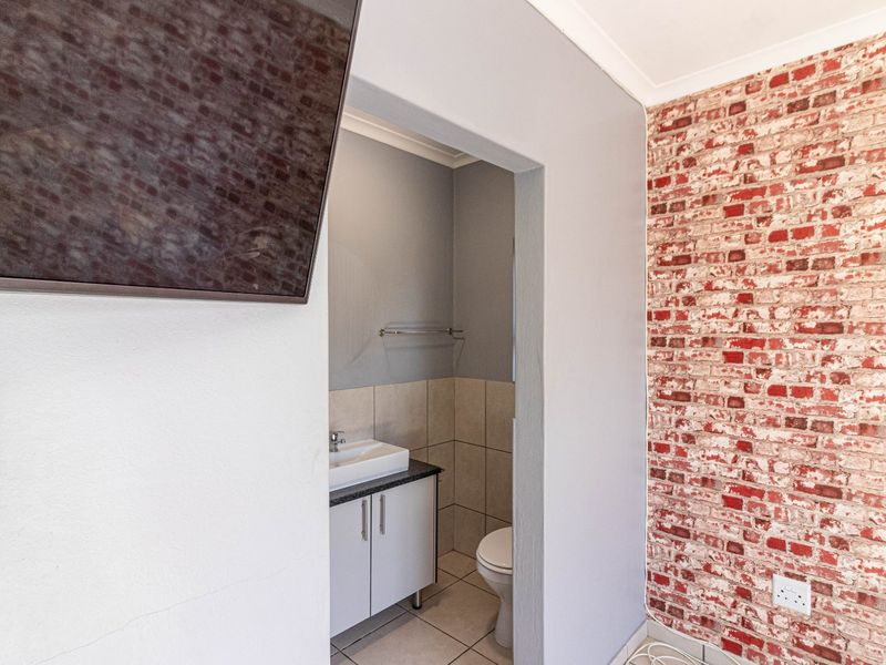 A compact bathroom featuring a vanity with basin, toilet, and tiled finishes, viewed from an adjacent room with a brick-effect feature wall.