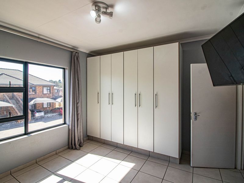 A bright, empty bedroom featuring neutral tiled flooring, built-in white cupboards, and large aluminium-framed windows providing natural light.