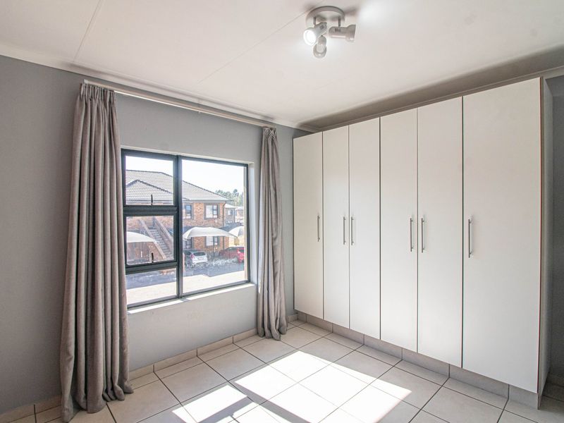 A bright, empty bedroom featuring neutral grey walls, tiled flooring, and extensive built-in white cupboards with modern hardware.
