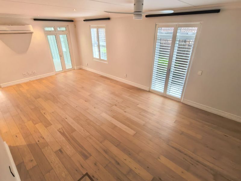 Spacious living area featuring light timber flooring, plantation shutters, ceiling fan, and split-system air conditioning with direct access to the exterior.