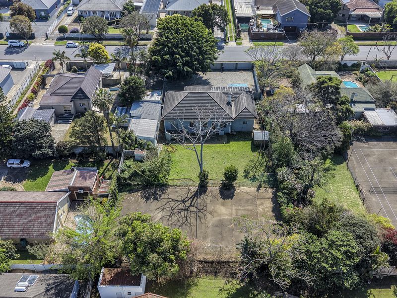 Aerial view of the property showcasing the residential dwelling, large garden, private tennis court, and swimming pool within a suburban neighborhood setting.