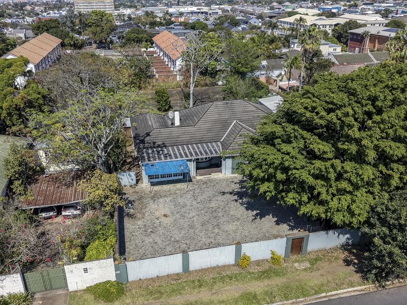 Aerial view of the property featuring a tiled roof house, large paved driveway, perimeter wall, and a detached carport structure.