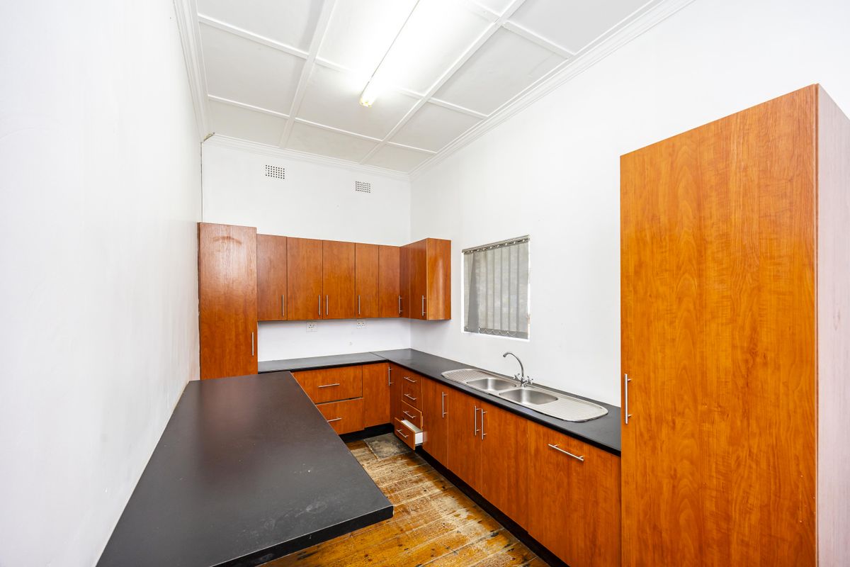 A functional kitchen featuring wooden cabinetry, dark laminate countertops, a stainless steel double sink, and original timber flooring.
