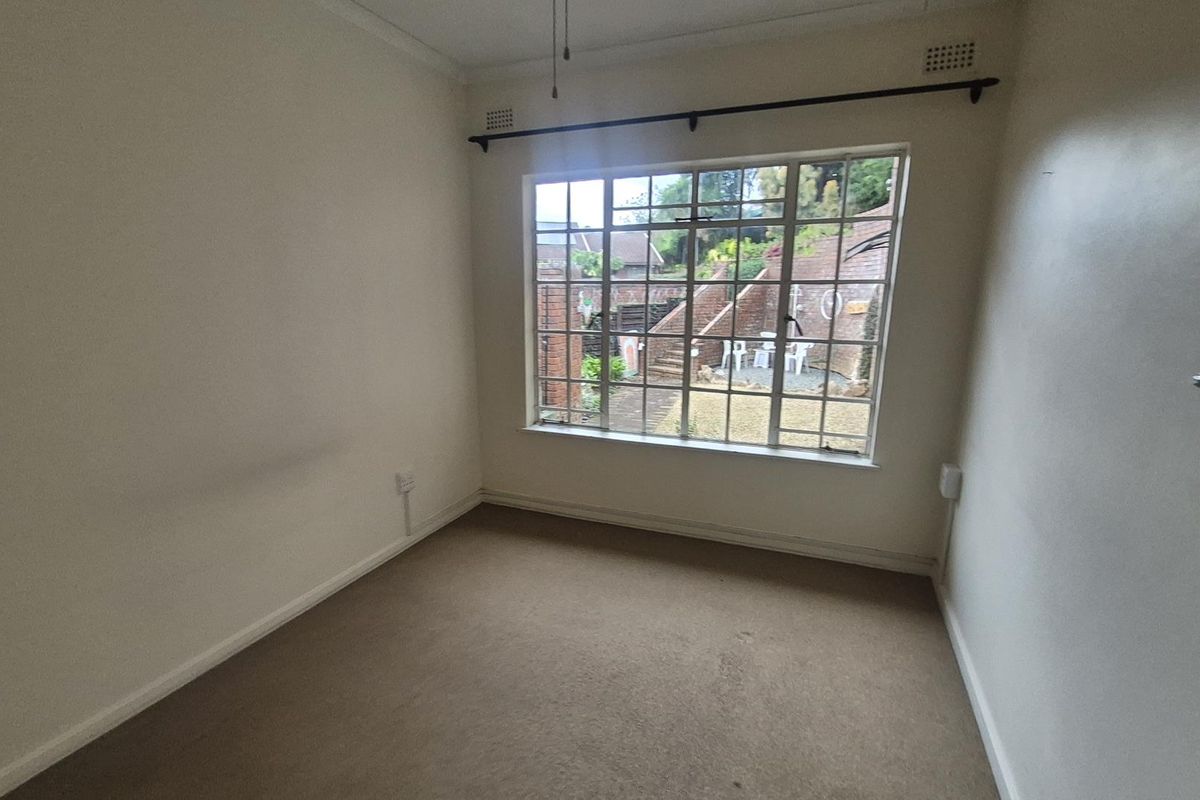 An empty bedroom featuring carpeted flooring and a large steel-framed window overlooking the rear garden area.