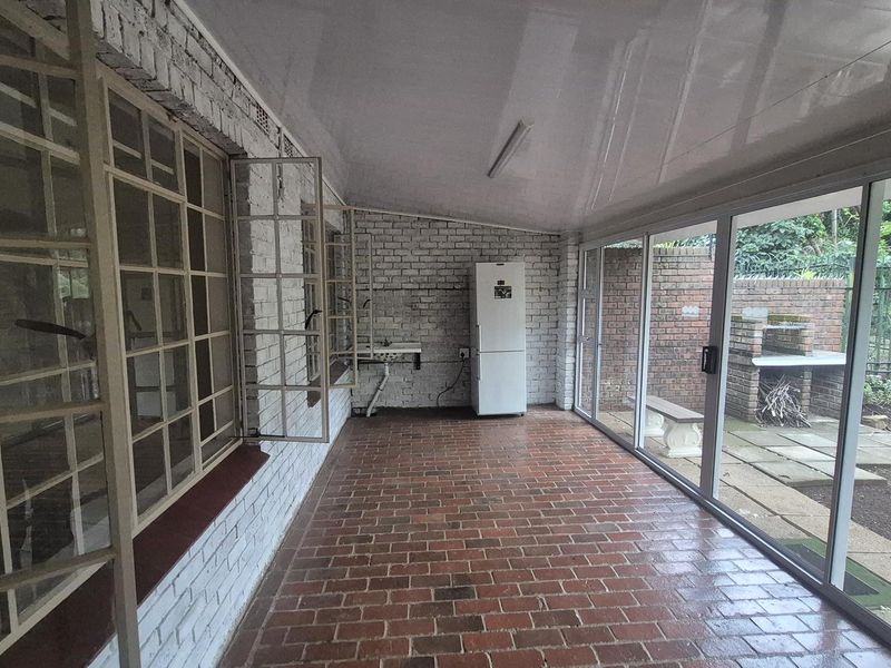 A spacious covered patio featuring brick flooring, exposed brick walls, and large sliding glass doors, providing seamless access to the outdoor garden and braai area.