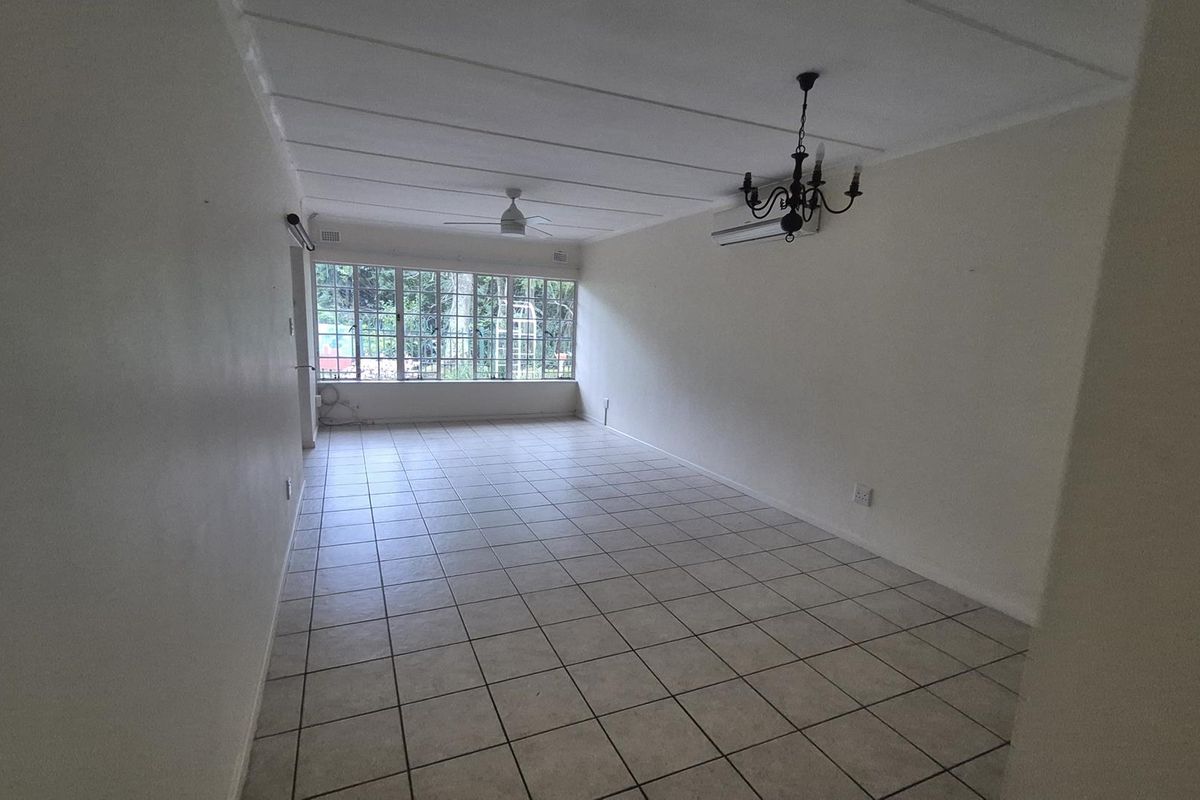 A spacious, empty living room featuring tiled flooring, a ceiling fan, a wall-mounted air conditioner, and large windows providing natural light.