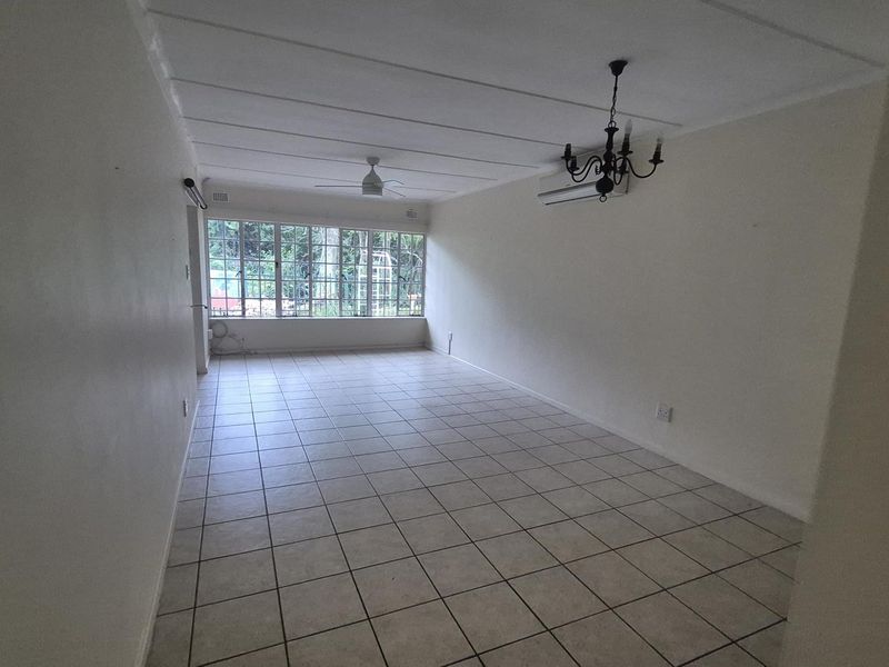 A spacious, empty living room featuring tiled flooring, a ceiling fan, a wall-mounted air conditioner, and large windows providing natural light.