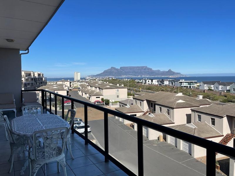 A spacious covered balcony featuring tiled flooring and a glass balustrade, offering expansive views of the surrounding neighborhood and Table Mountain.
