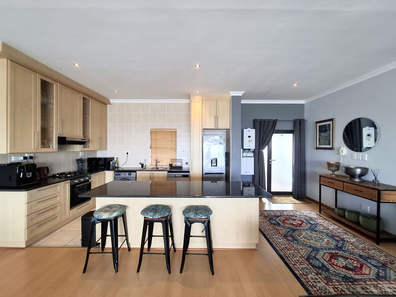 Spacious open-plan kitchen featuring light wood cabinetry, a large granite island, built-in appliances, and recessed lighting, seamlessly flowing into the living area.