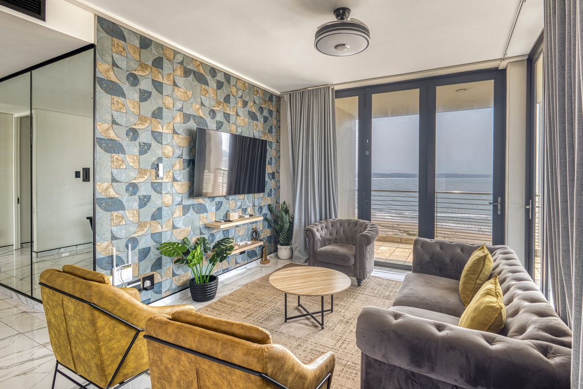 Modern living room featuring a striking patterned feature wall, marble-look flooring, and large glass doors opening to a balcony with ocean views.