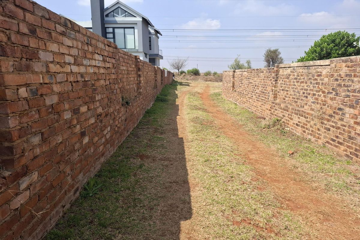 A view of a vacant residential plot featuring long brick boundary walls and an unpaved access path leading towards the rear of the property.