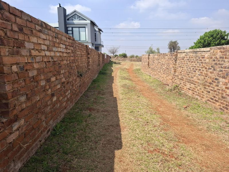 A view of a vacant residential plot featuring long brick boundary walls and an unpaved access path leading towards the rear of the property.