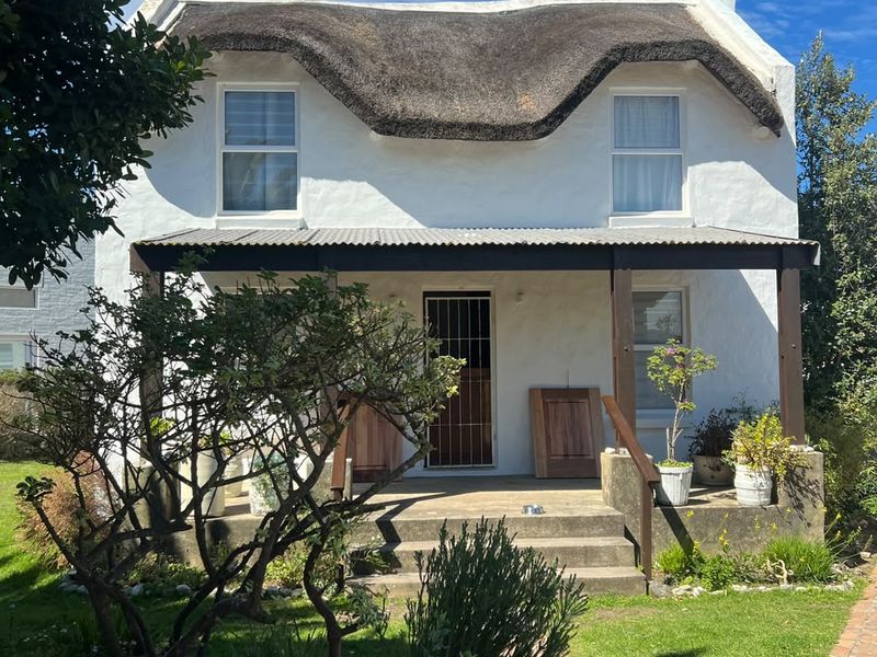 Charming double-story cottage featuring a traditional thatched roof, a covered front veranda, and a well-established garden entrance.