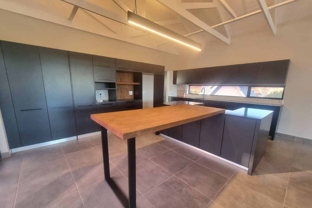 Modern kitchen featuring dark cabinetry, a timber-topped island, tiled flooring, and exposed white roof trusses, creating a spacious and contemporary culinary environment.