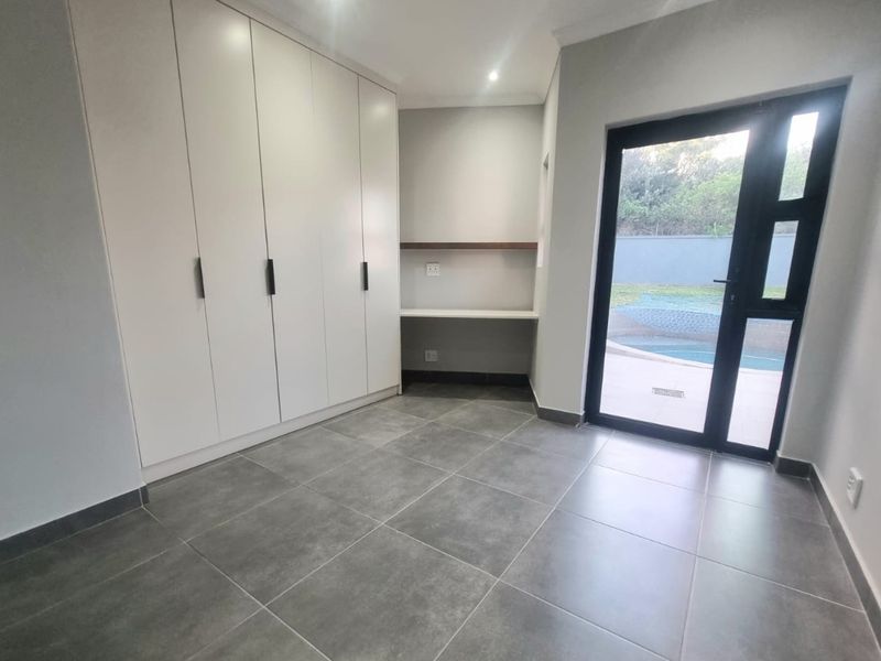 A bright, modern home office featuring sleek built-in cupboards, a dedicated desk area, tiled flooring, and direct access to the outdoor pool area.