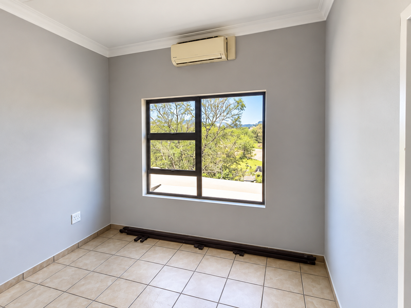 A bright, empty bedroom featuring tiled flooring, a wall-mounted air conditioning unit, and a large window with views of the surrounding greenery.