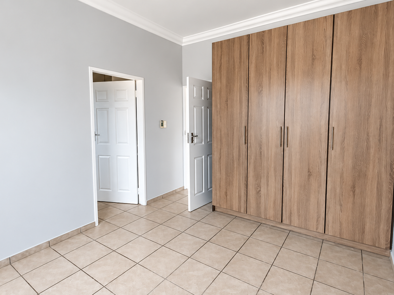 A bright, empty bedroom featuring neutral walls, tiled flooring, and a large, modern wood-finish built-in wardrobe.