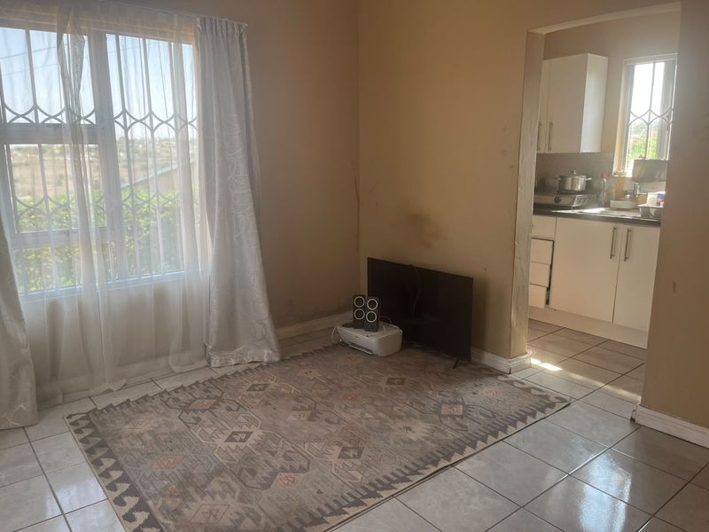 Open plan living area with tiled flooring and adjacent kitchen featuring built-in cabinetry and sink, showing signs of wall wear and staining.