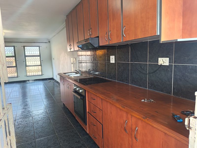 A functional kitchen featuring wooden cabinetry, built-in oven and hob, tiled backsplash, and dark floor tiles, leading into an open-plan living area.