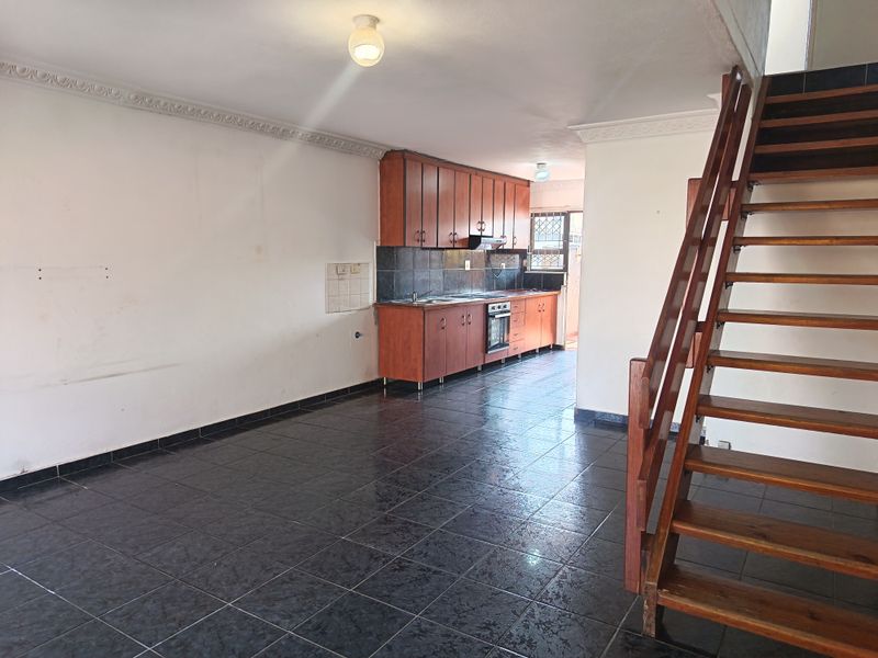 Open-plan living area featuring dark tiled flooring, a functional kitchen with built-in cabinetry, and a wooden staircase leading to the upper level.