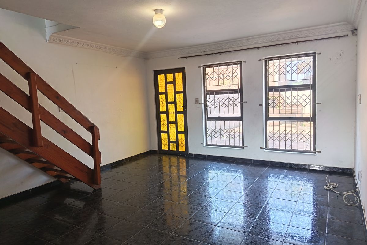 An empty living room featuring dark tiled flooring, a wooden staircase, and security-fitted windows and door.