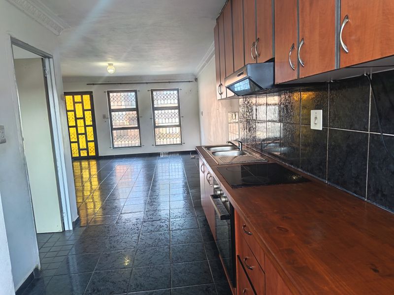 Open-plan kitchen and living area featuring dark tiled flooring, wooden cabinetry, built-in oven and hob, and security bars on the windows.