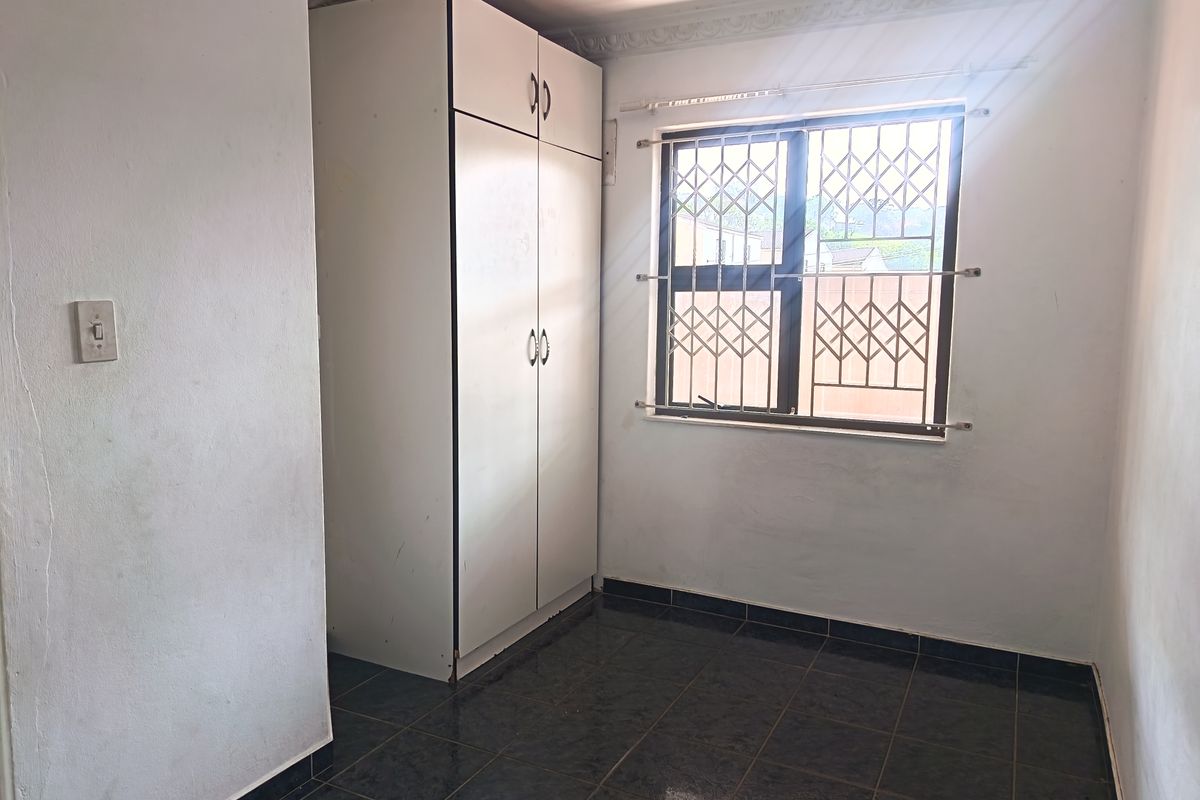 A compact, empty bedroom featuring dark tiled flooring, a built-in wardrobe, and a window fitted with a security gate.