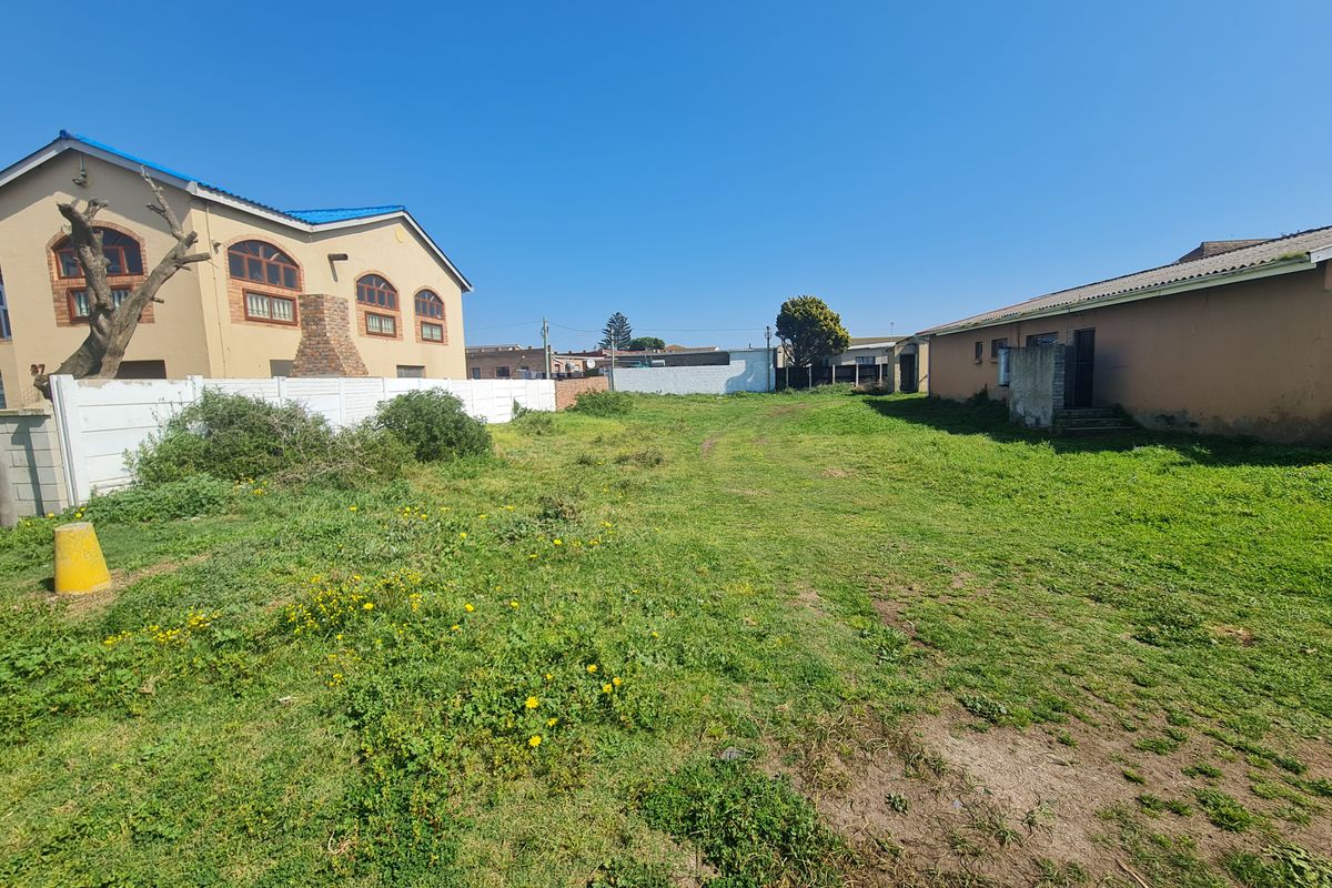 A vacant residential plot featuring open grassed space, enclosed by boundary walls and adjacent to existing residential structures.
