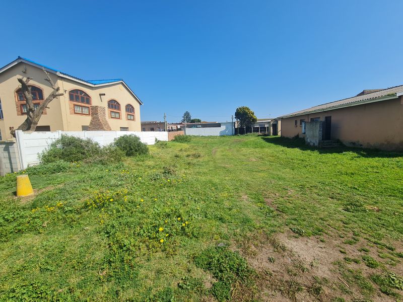 A vacant residential plot featuring open grassed space, enclosed by boundary walls and adjacent to existing residential structures.