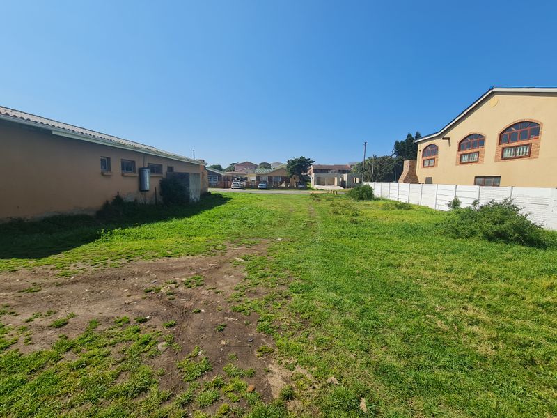 A vacant residential plot featuring a grass lawn, dirt driveway, and precast concrete boundary wall, situated between existing neighboring properties.