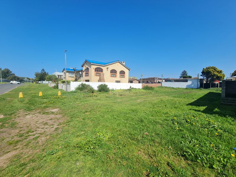 A view of a vacant residential plot featuring a grass lawn and boundary wall, with neighboring properties visible in the background.