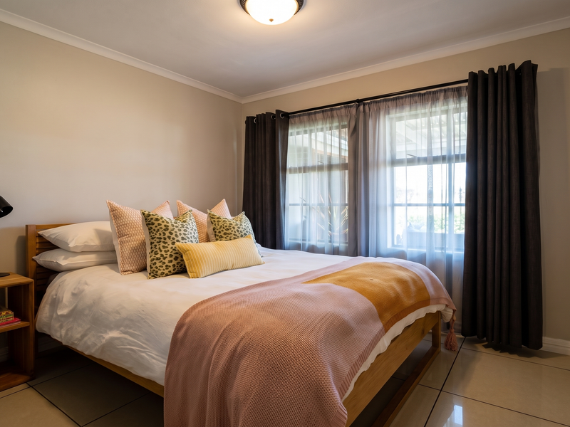 A well-maintained bedroom featuring neutral tiled flooring, ample built-in cupboard storage, and natural light through a window with security bars.