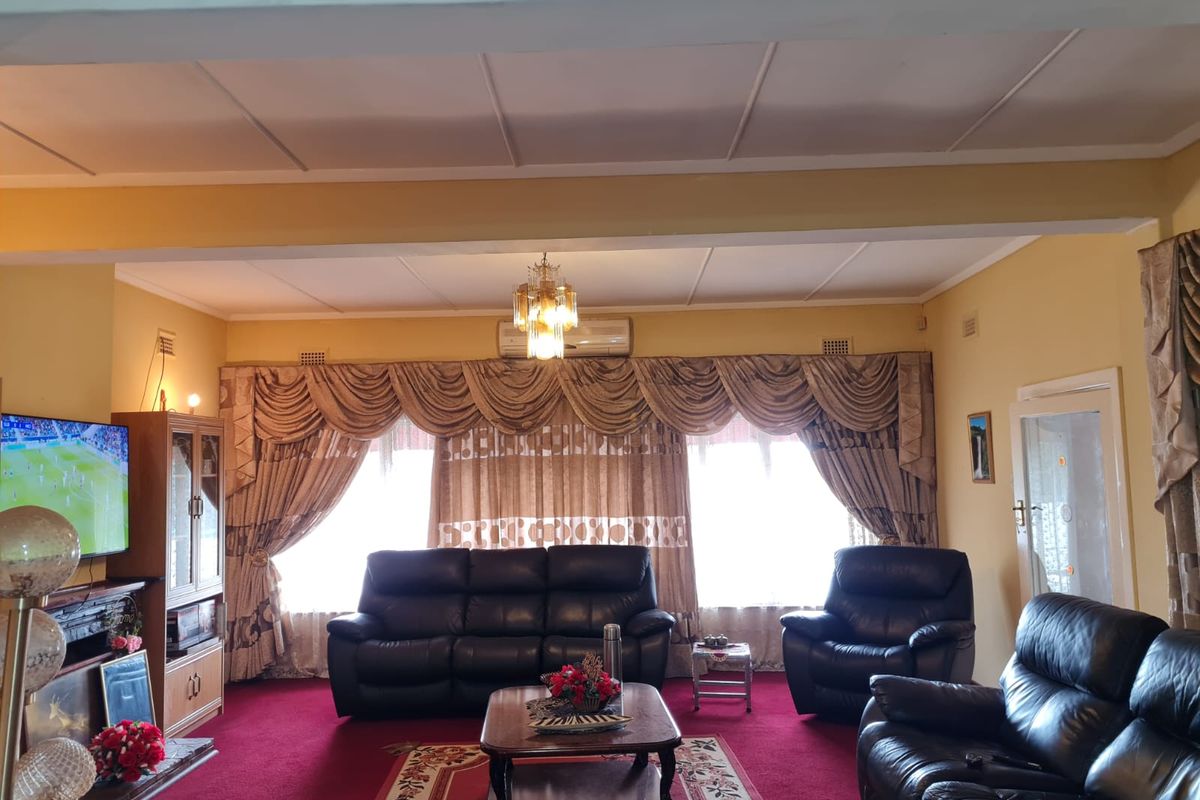 A traditional living room featuring a central chandelier, wall-mounted air conditioning unit, and built-in cabinetry, with carpeted flooring and large window treatments.