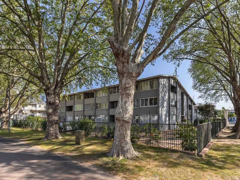 Exterior view of a multi-story apartment building featuring a grey facade, surrounded by mature trees and secured by metal perimeter fencing.