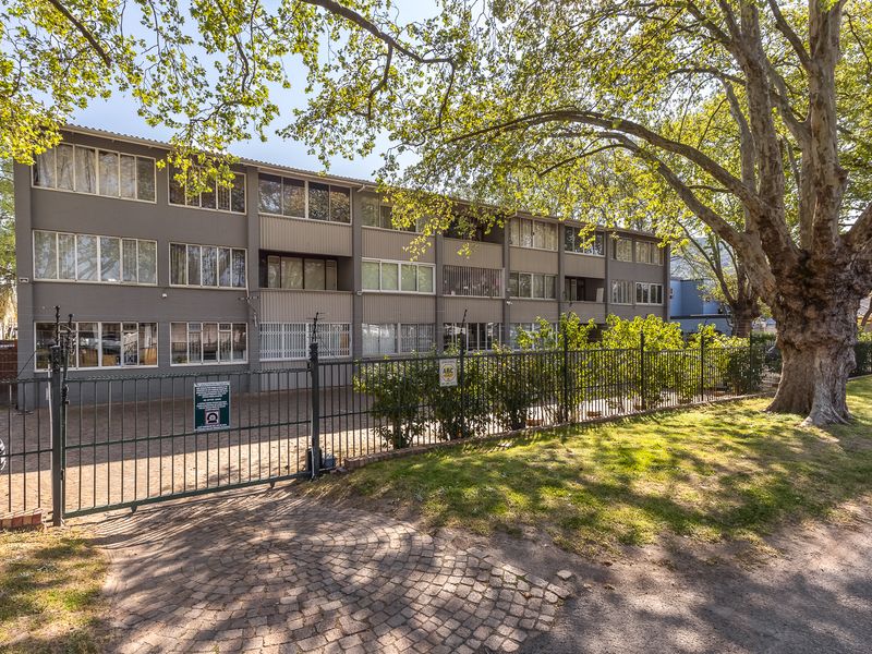 Exterior view of a multi-story apartment building featuring a paved driveway, secure metal fencing, and mature trees in a well-established residential setting.