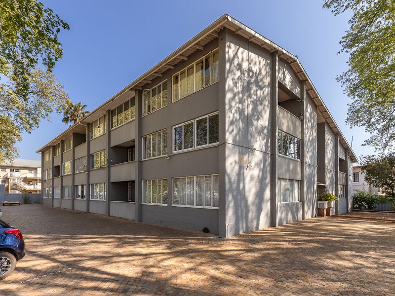 A multi-story apartment building featuring a grey painted brick facade, paved driveway, and multiple levels of balconies and casement windows.