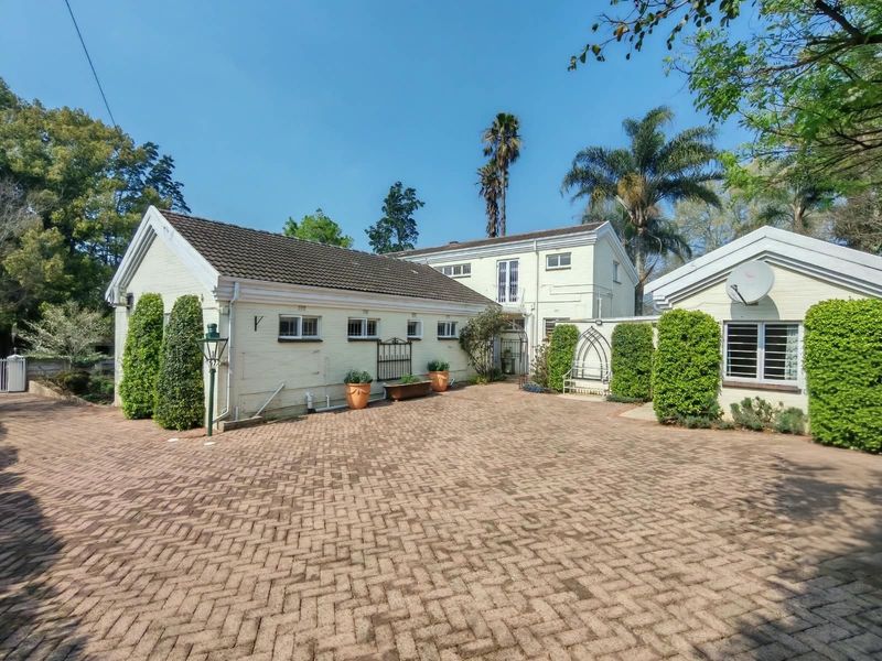 Front exterior view of the property featuring a large brick-paved driveway, painted brick facade, and established garden surroundings.