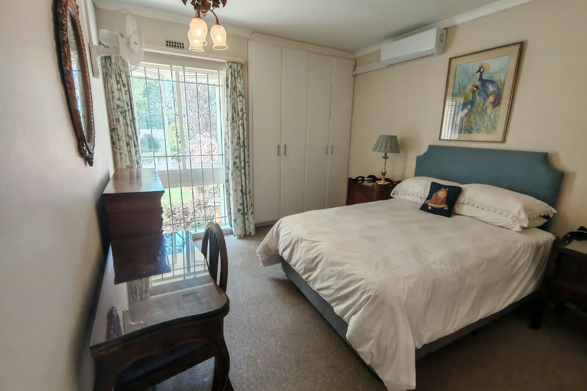 A functional bedroom featuring built-in cupboards, carpeted flooring, and a wall-mounted air conditioning unit, with natural light provided by a window with security bars.