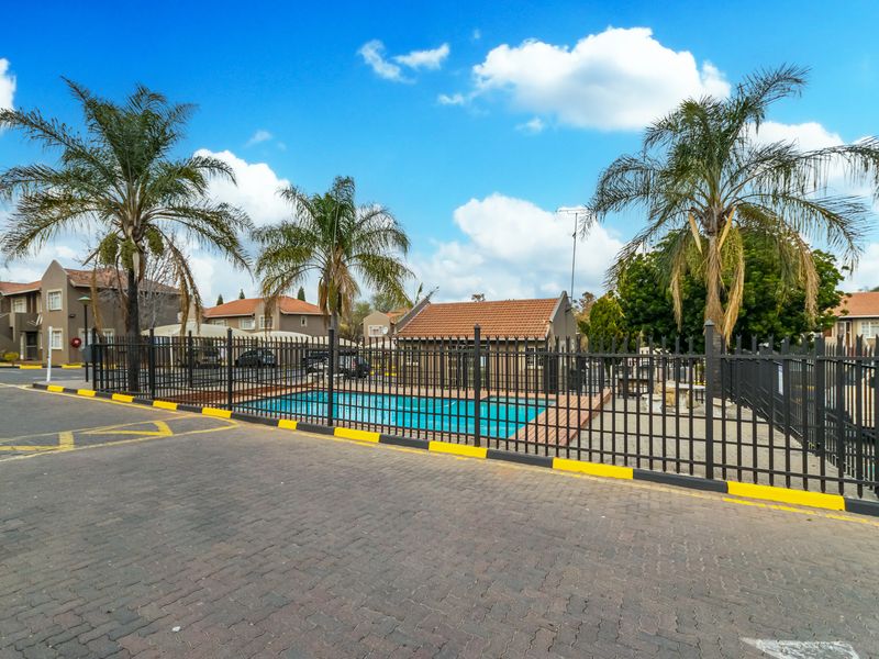 A communal swimming pool area featuring secure metal perimeter fencing, paved surroundings, and established palm trees within an apartment complex.