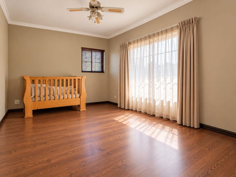 A spacious, well-lit bedroom featuring warm-toned laminate flooring, neutral wall paint, a ceiling fan, and large windows dressed with curtains and blinds.