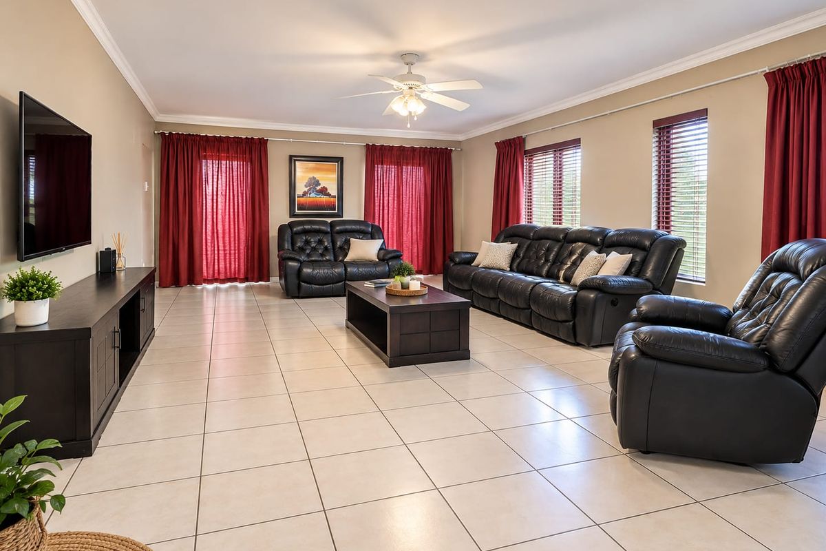 Spacious living room featuring neutral tiled flooring, crown molding, a central ceiling fan, and large windows with venetian blinds.