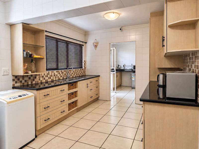 A functional kitchen featuring light wood cabinetry, granite countertops, tiled backsplash, and tiled flooring, with an adjoining scullery area visible through the doorway.