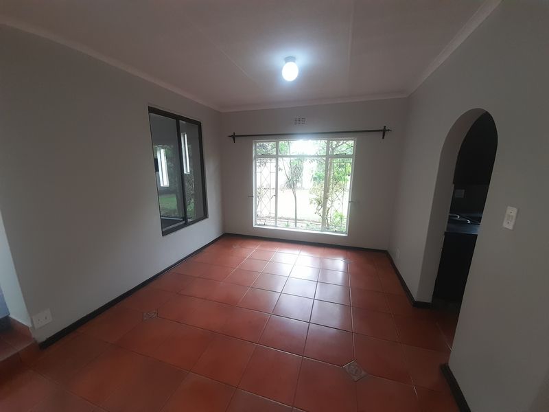 An empty living room featuring terracotta-style floor tiles, an arched entryway, and large windows with security bars overlooking the garden.
