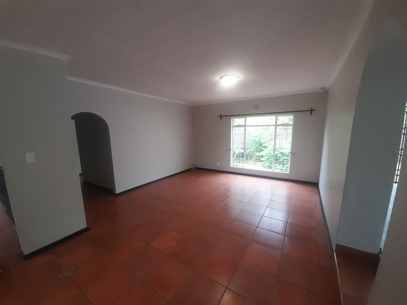 A spacious, empty living room featuring terracotta floor tiles, an arched entryway, and a large window with security bars.