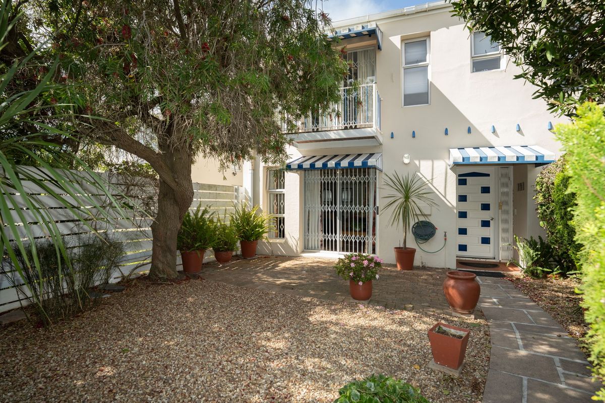 Rear exterior view featuring a paved patio area, gravel garden, security gate, and a balcony with striped awnings.
