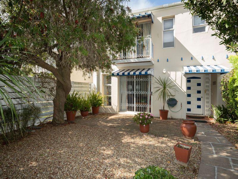 Rear exterior view featuring a paved patio area, gravel garden, security gate, and a balcony with striped awnings.