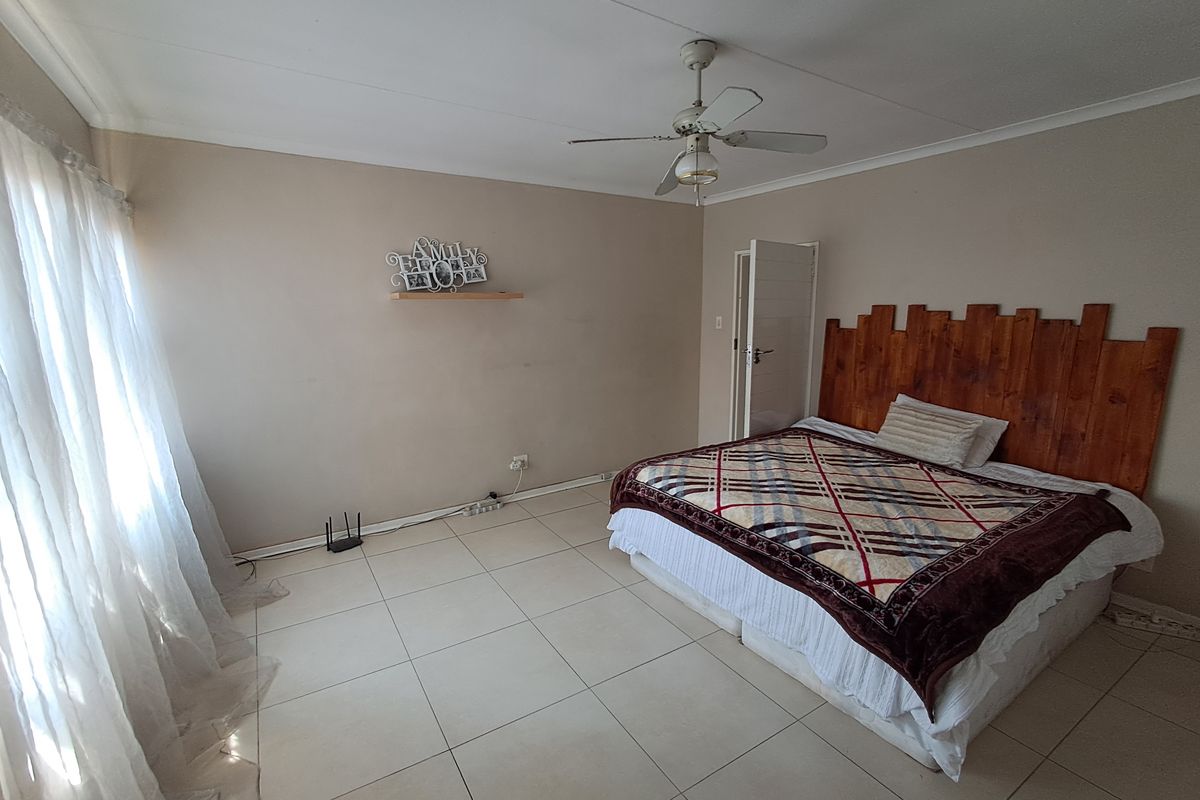 A spacious bedroom featuring neutral tiled flooring, a ceiling fan, and a wooden headboard feature wall.
