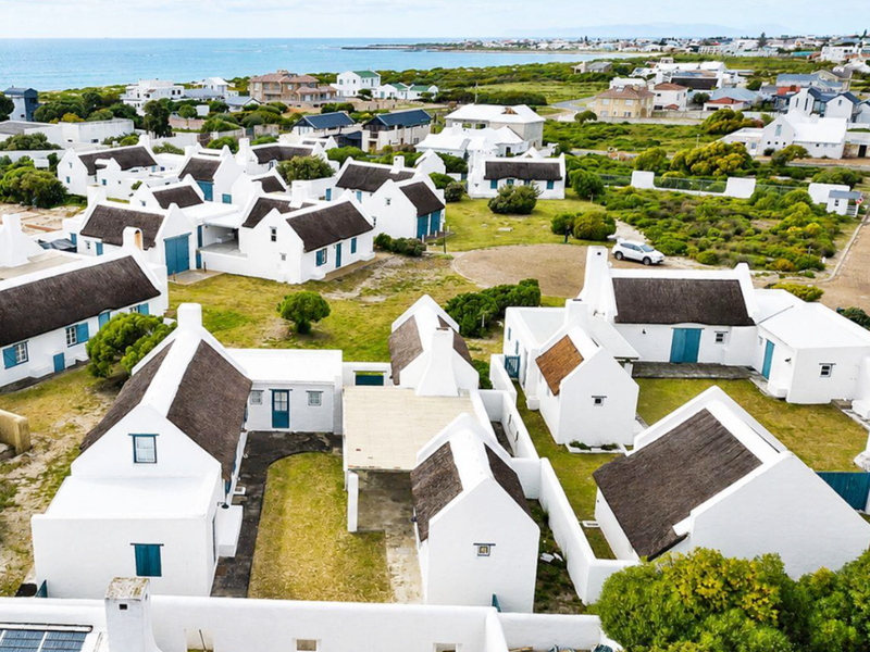 An aerial view of a coastal residential estate featuring multiple white cottages with traditional thatched roofs and surrounding landscaped grounds.